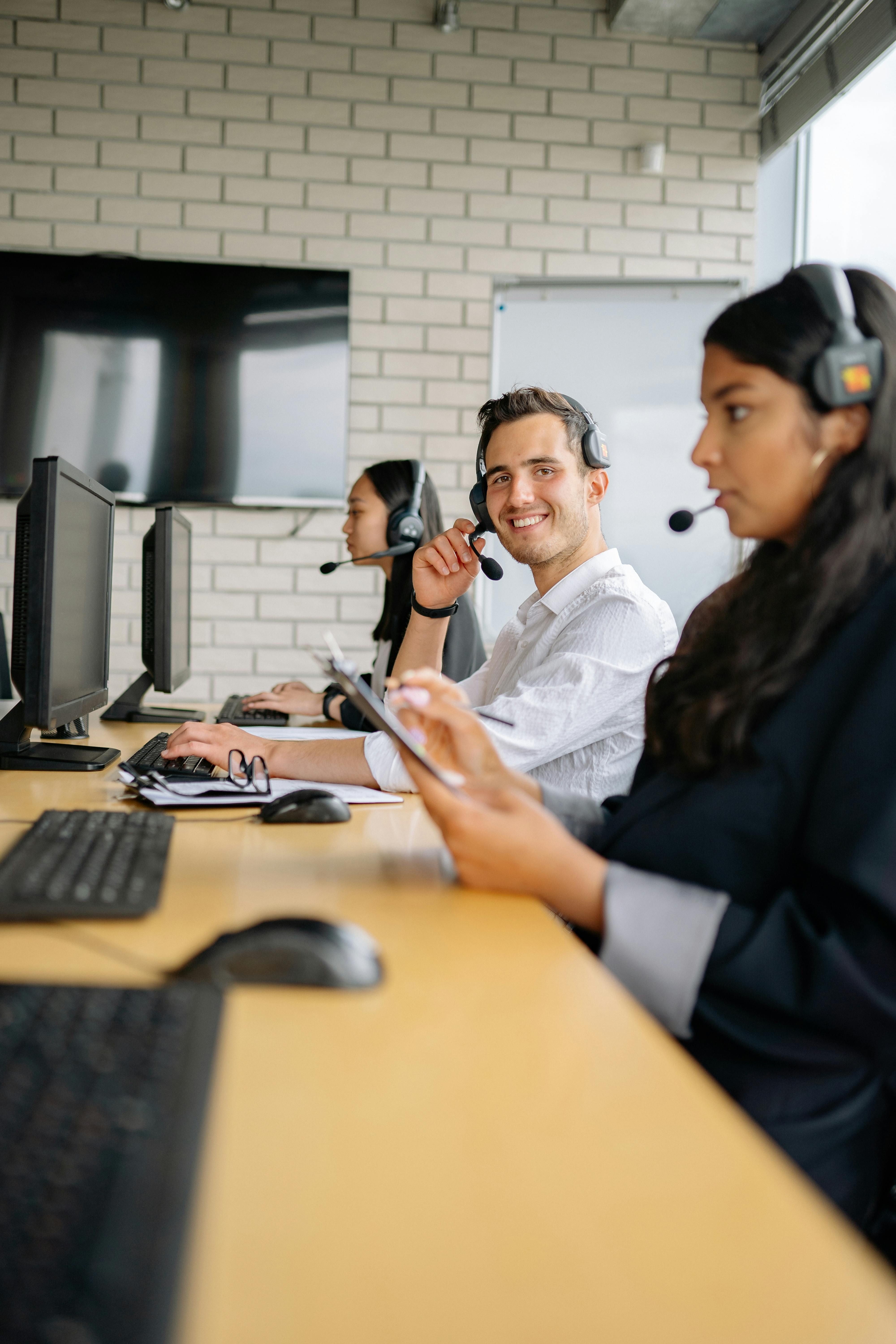 Three call center agents wearing headsets, engaged in conversations at their desks in a busy office environment.